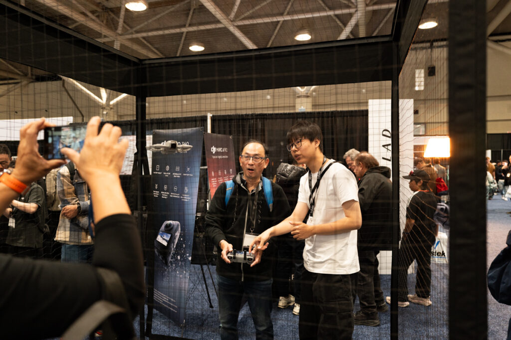 Two men are flying a drone in a netted cage at the DJI booth at ProFusion Expo 2025 in Toronto.